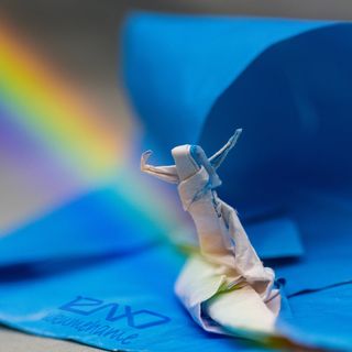 A white origami surfer and a blue origami wave. A rainbow ends at the surfer's feet.